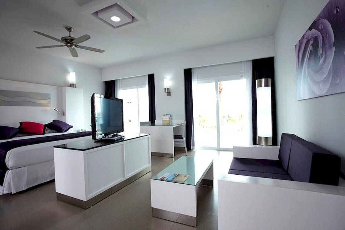 Innenansicht einer Suite im Riu Palace Peninsula Hotel mit Bett, Fernseher, Couch und Schreibtisch.
