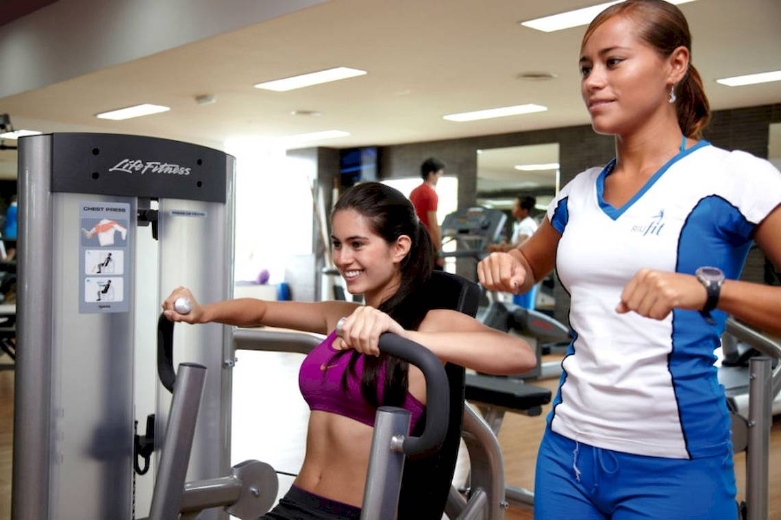 Frau im Fitnessstudio trainiert mit einer Trainerin auf einer Brustpresse-Maschine.