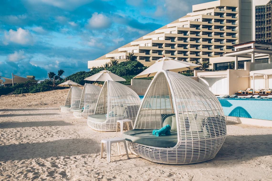 Strandbereich des Coral Level at Iberostar Selection Cancun mit weißen Rattan-Loungesesseln und Sonnenschirmen.