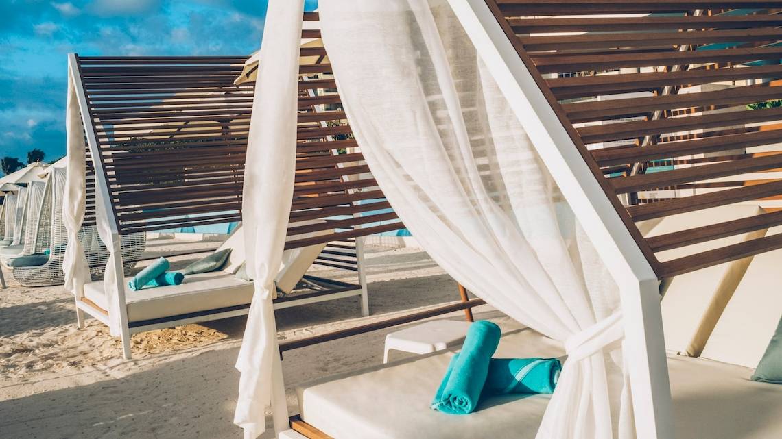 Reihe von Loungebetten am Strand des Coral Level at Iberostar Selection Cancun, bedeckt mit weißen Vorhängen und blauen Handtüchern.