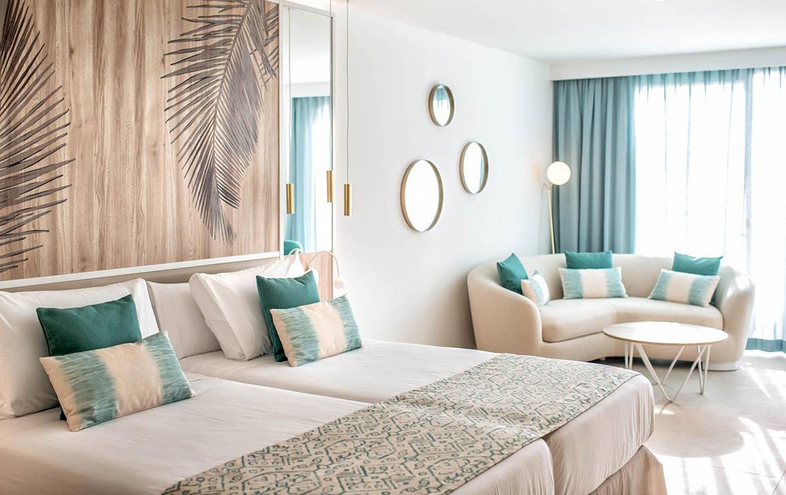 Ein modernes Hotelzimmer im Guayarmina Princess Hotel mit zwei Betten, blauen Kissen und einer gemütlichen Couch.