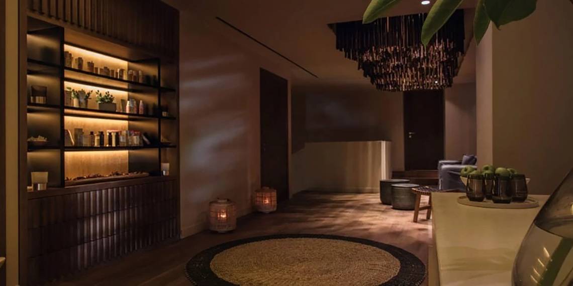 Innenansicht des Alion Beach Hotels mit beleuchteten Regalen, einem runden Teppich und einer geometrischen Deckenleuchte.