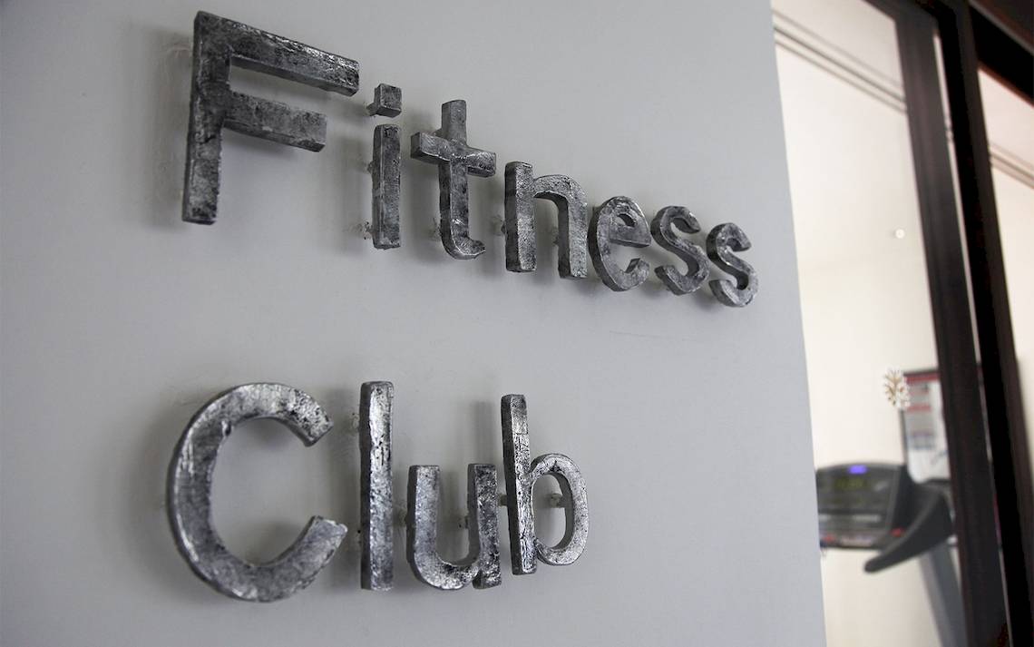 Schild mit den Worten 'Fitness Club' an einer Wand, mit einem Blick auf eine Tretmühle im Hintergrund.