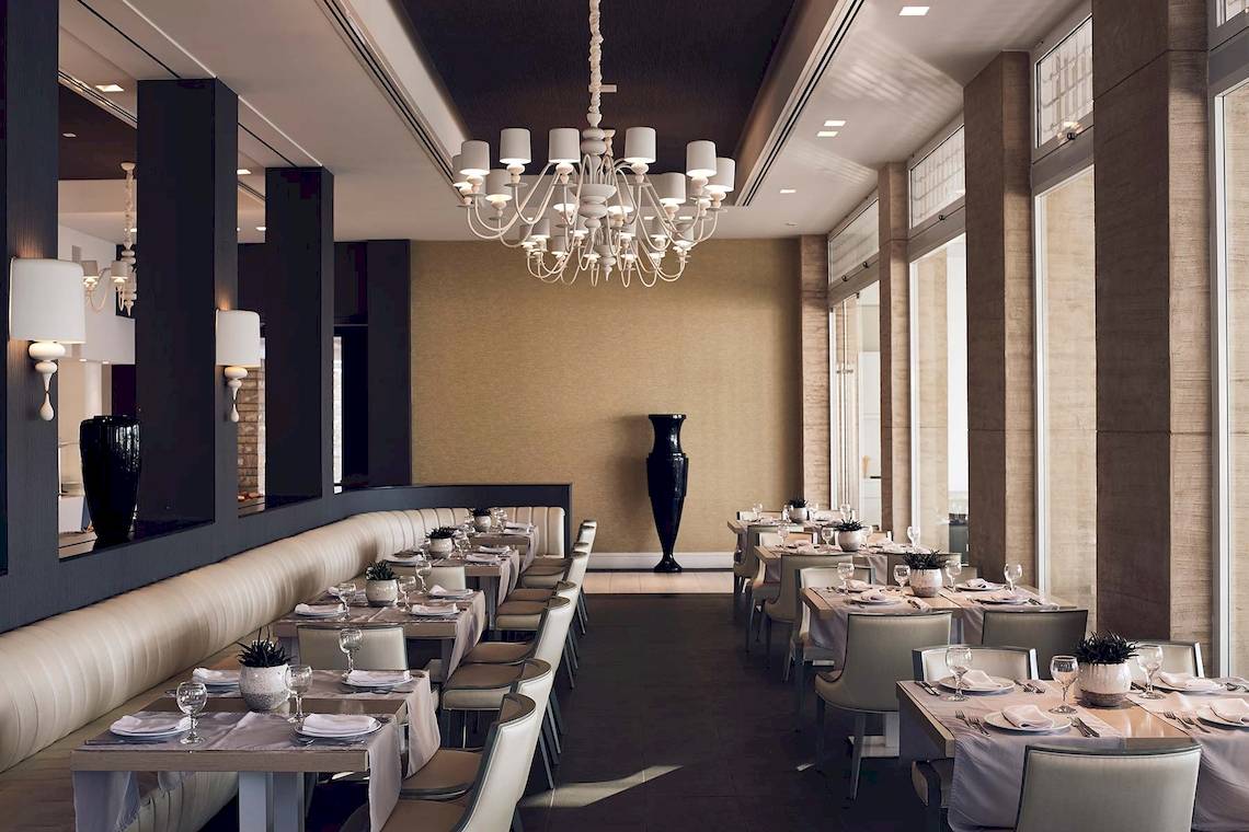 Innenbereich des Pelagos Suites Hotel & Spa mit elegant eingerichteten Tischen und Stühlen, großen Fenstern und einer beeindruckenden Kronleuchter.