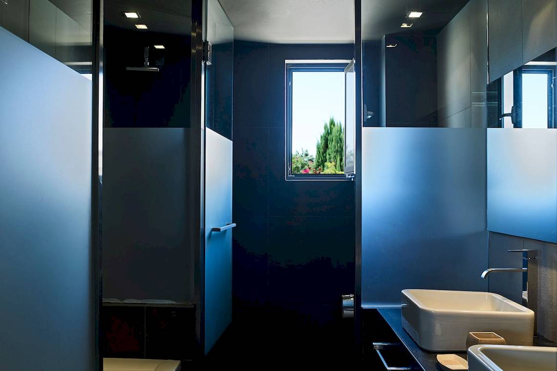 Badezimmer im Pelagos Suites Hotel & Spa mit blauen Fliesen, Dusche und offenem Fenster mit Gartenblick.