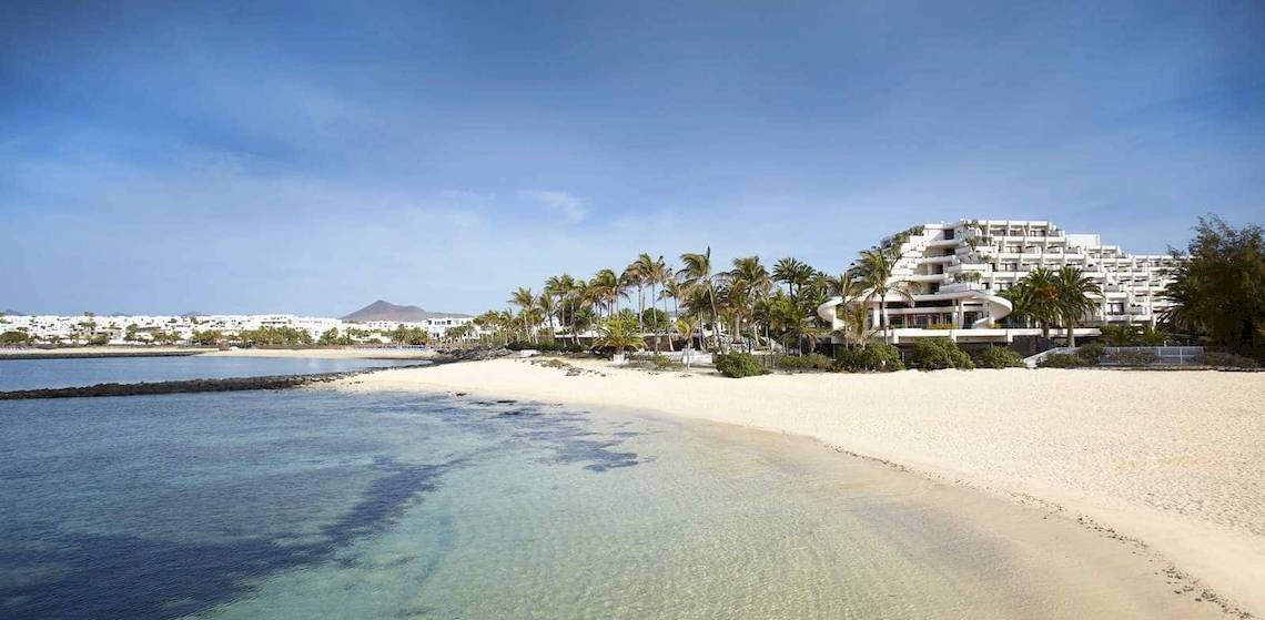 Außenansicht des Melia Salinas Hotels an einem weißen Sandstrand mit Palmen und Gebäuden im Hintergrund.