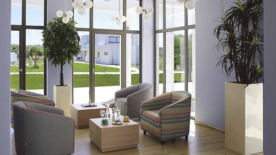 Lobby des Club Plimmiri Hotels mit großen Fenstern, modernen Möbeln und Pflanzen.