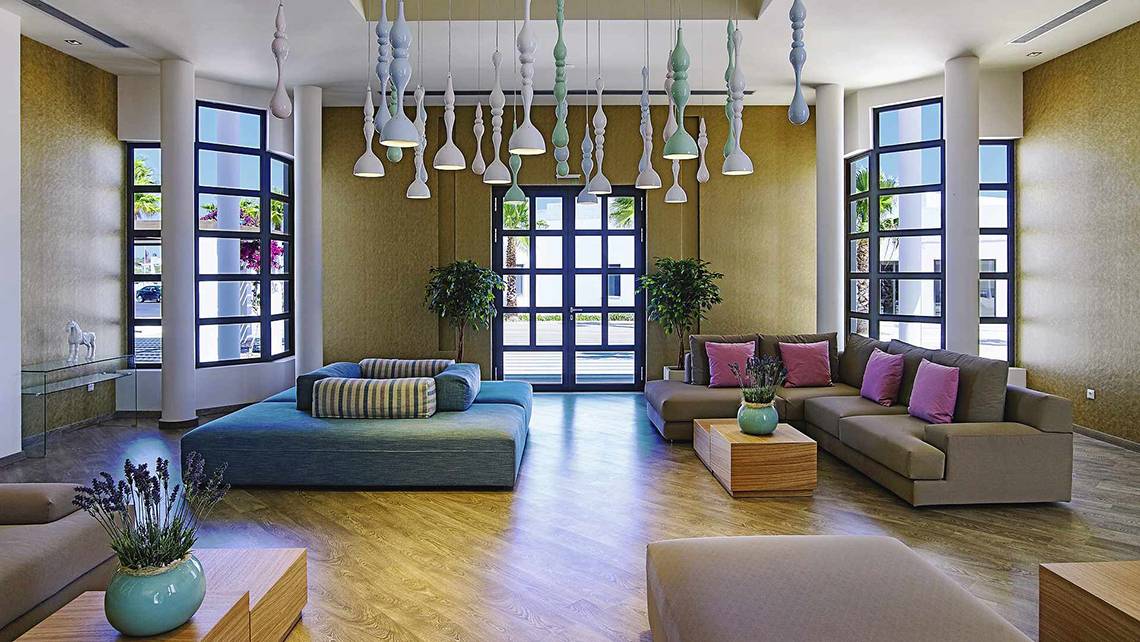 Lobby des Club Plimmiri Hotels mit modernen Möbeln, Holzboden und großen Fenstern.