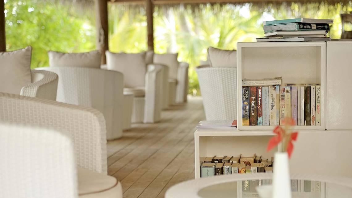 Bibliothek im Outrigger Maldives Maafushivaru Resort mit Büchern, Sesseln und Glastisch.