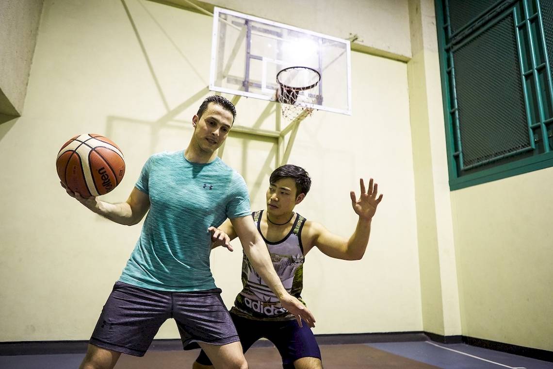 Zwei Männer spielen Basketball in einer Halle, einer hält den Ball und der andere versucht ihn zu blockieren.