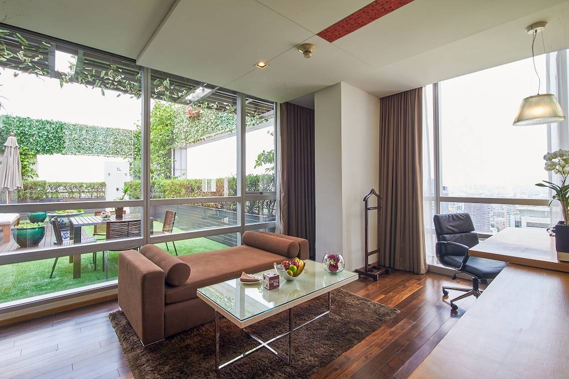 Innenansicht des Wohnbereichs im Pathumwan Princess Hotel mit großem Fenster, Gartenblick und moderner Einrichtung.