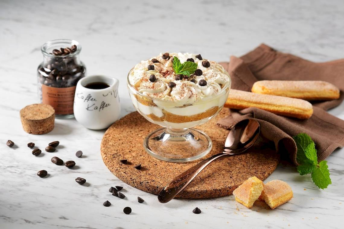 Ein Tiramisu-Dessert auf einem Tisch mit Kaffee, Keksen und Minzeblättern.