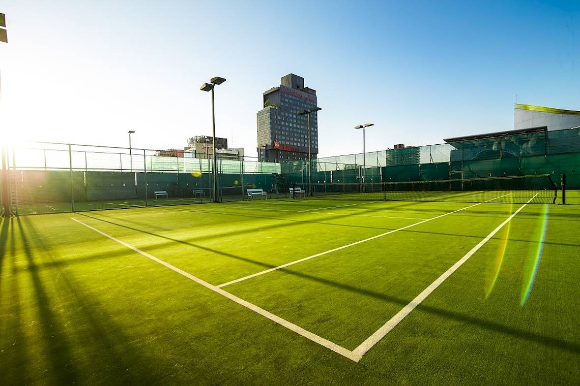 Tennisplatz mit Zäunen und Gebäuden im Hintergrund, alles in hellem Sonnenlicht