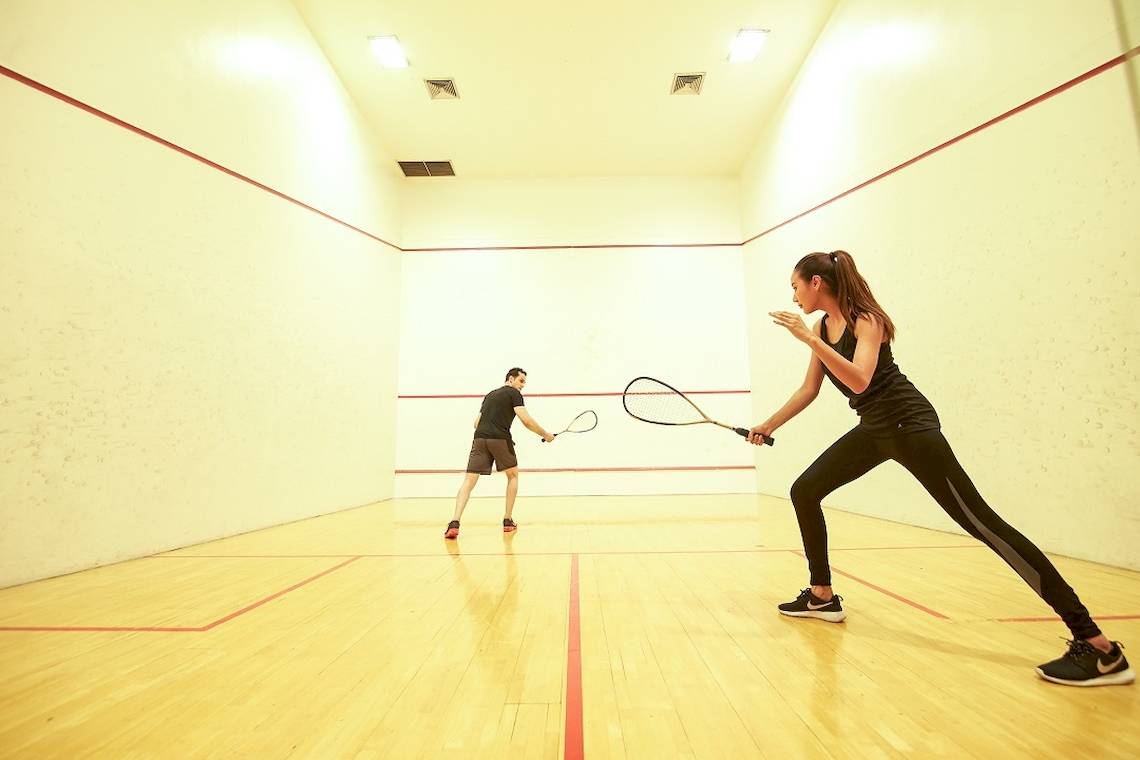Zwei Personen spielen Squash in einem Innenraum mit Holzboden und roten Linien.