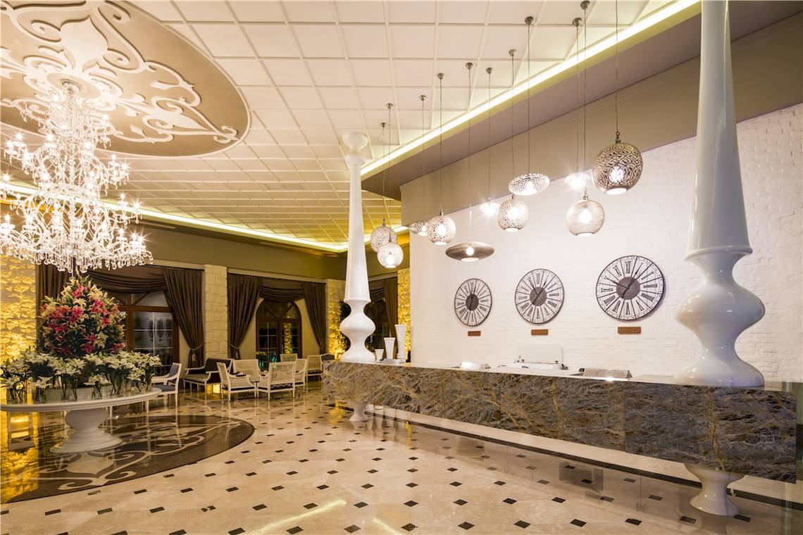 Empfangsbereich des Voyage Torba Hotels mit Marmorrezeption, Kronleuchter und Uhren an der Wand.