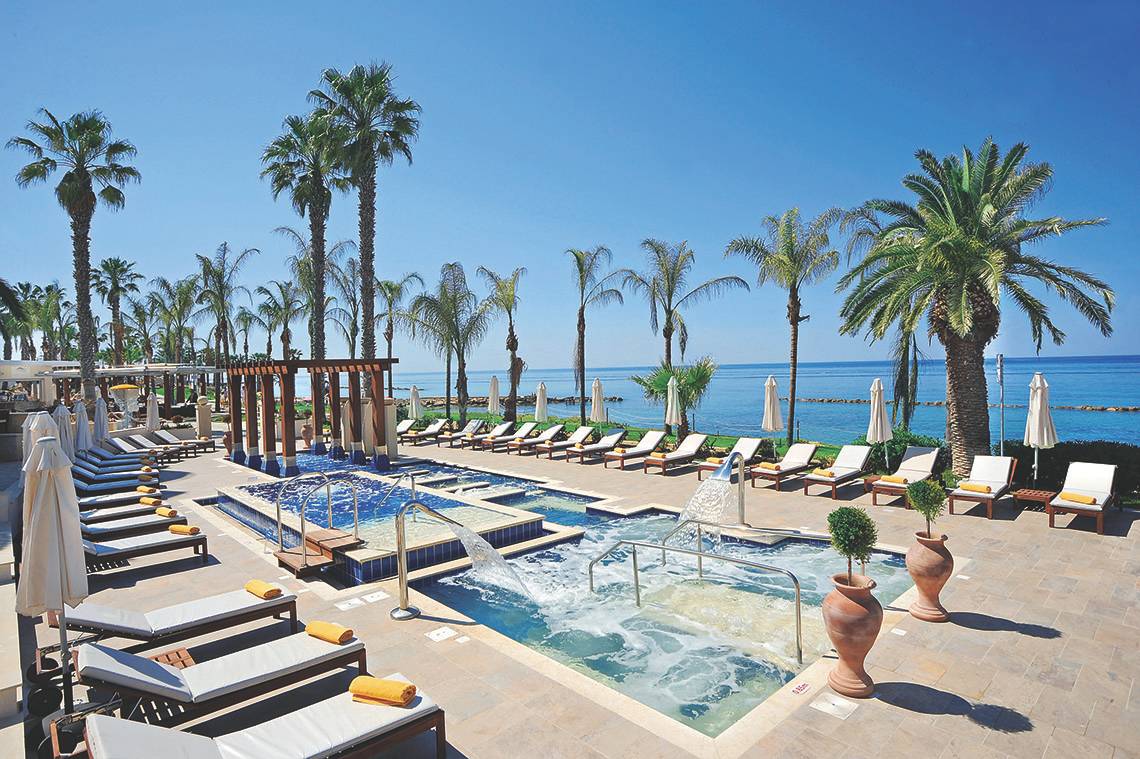 Außenpoolbereich des Alexander The Great Hotels mit Loungeliegen, Palmen und Meerblick.