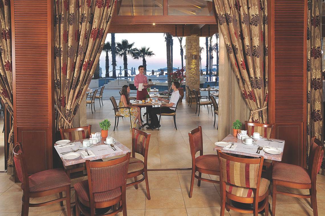 Innenansicht des Restaurants im Alexander The Great Hotel mit Gästen und Palmen im Hintergrund.