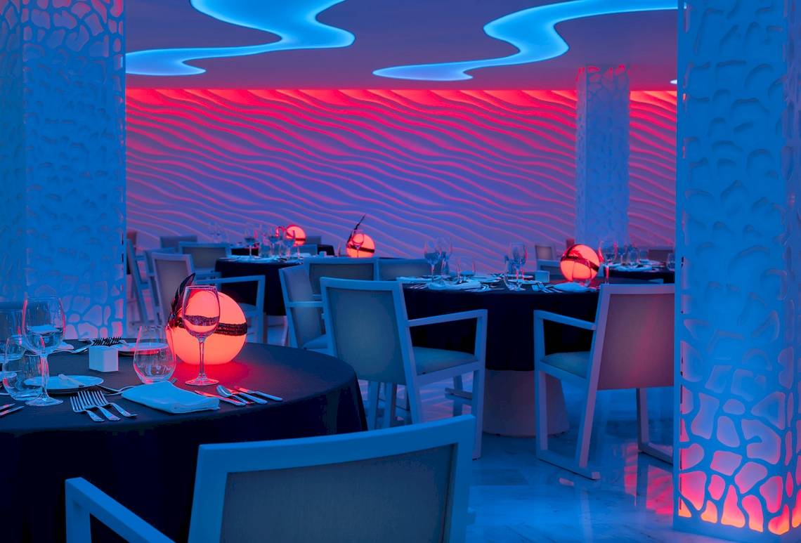 Innenraum des H10 Conquistador Hotels mit blauen und roten Lichtern und gedeckten Tischen.