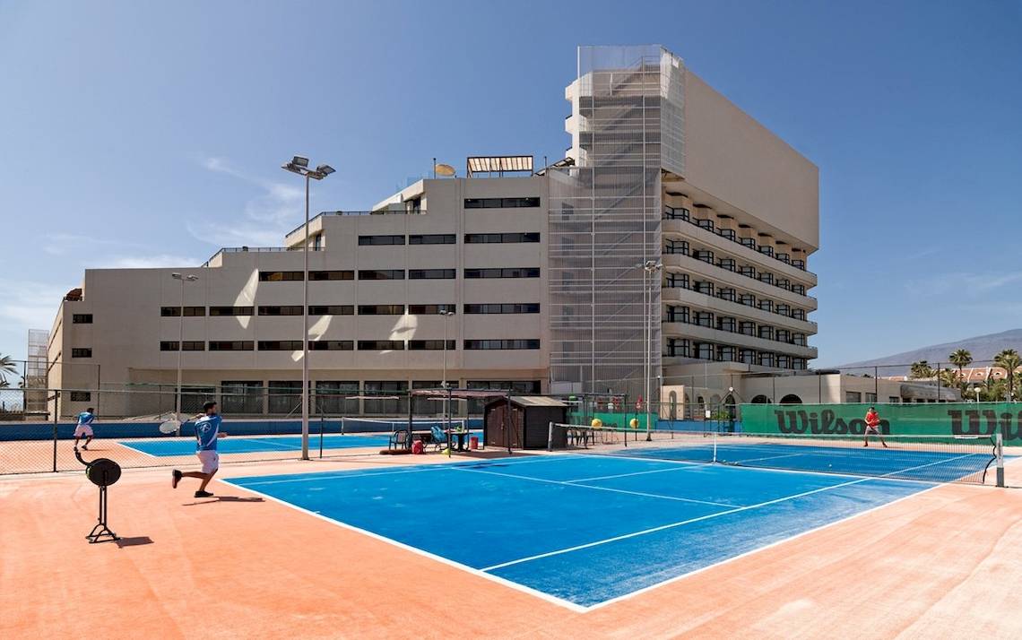 Tennisplatz vor dem H10 Conquistador Hotel mit Spielern und Gebäude im Hintergrund.