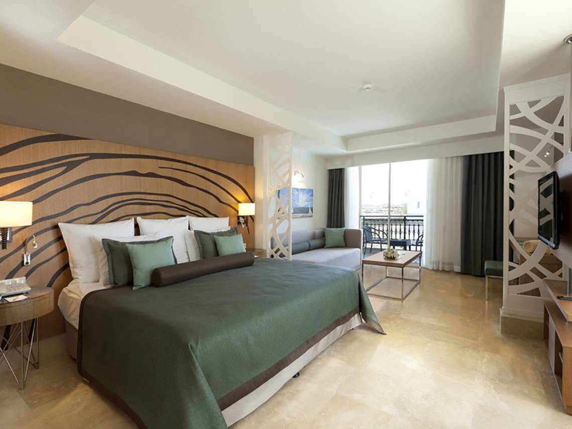 Innenansicht eines eleganten Hotelzimmers im Paloma Oceana Hotel mit Bett, Wohnzimmerbereich und Terrasse.