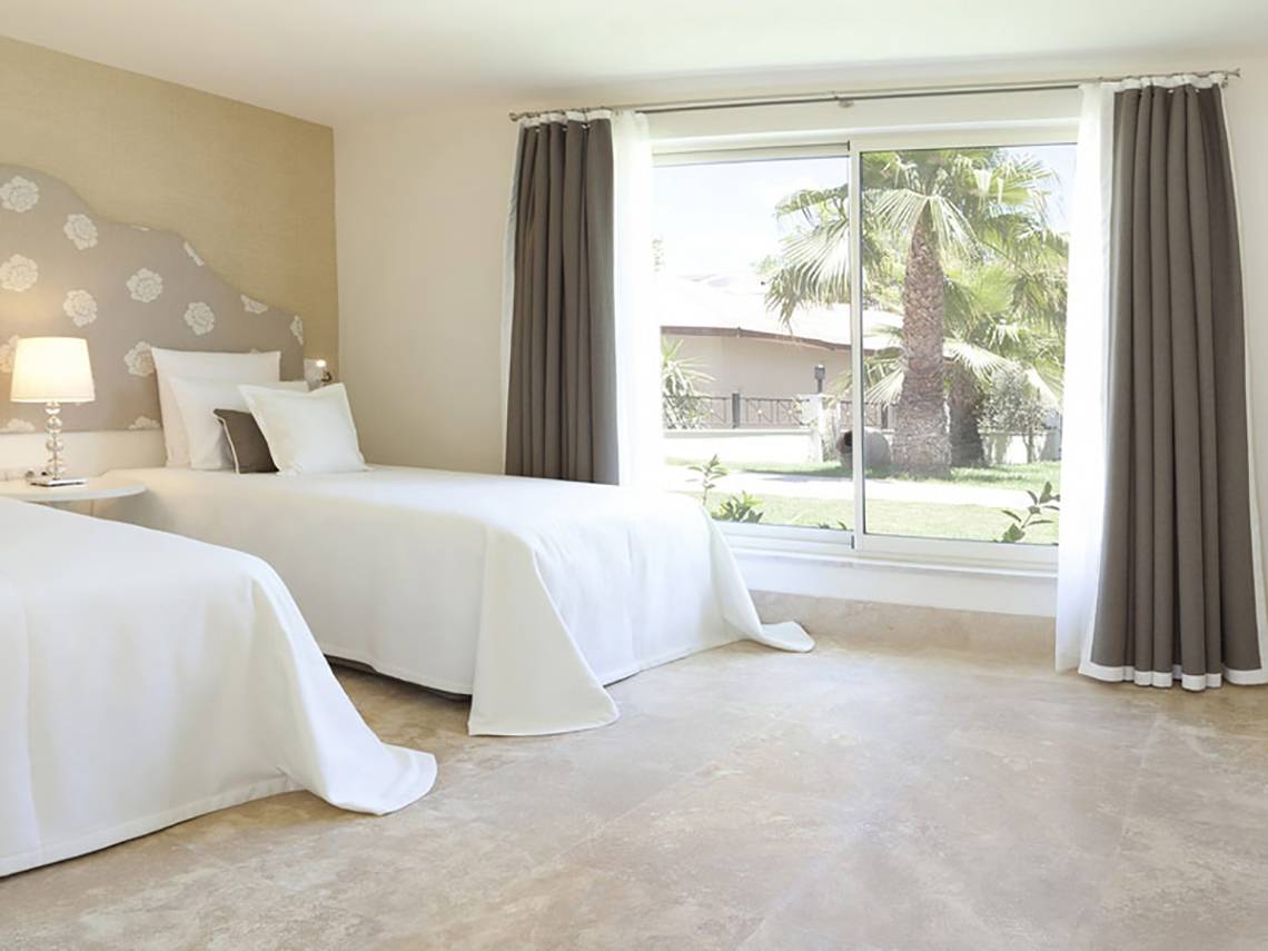 Zwei Betten im Zimmer des Paloma Oceana Hotels mit weißem Laken und Kissen, neben einem Fenster mit Blick auf einen Garten.