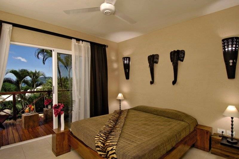 Schlafzimmer mit Aussicht auf den Balkon und Palmen, komplett eingerichtet mit Bett, Lampen und Elefanten-Wandleuchten.