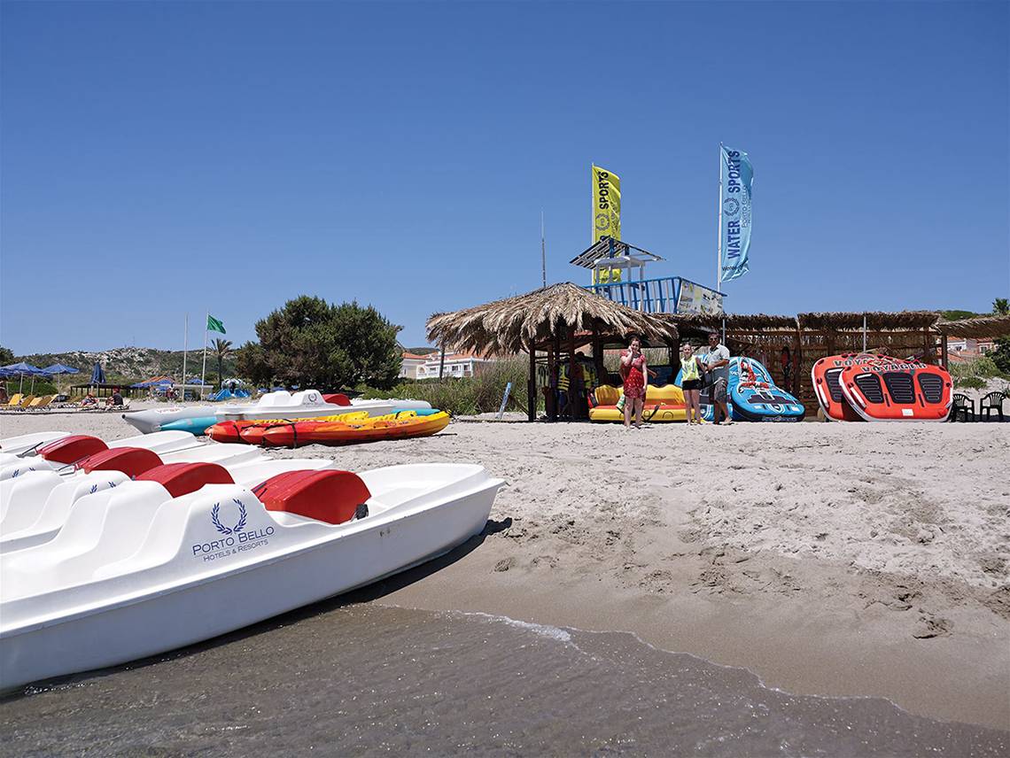 Strand des Porto Bello Hotels mit mehreren Booten und Gästen in der Nähe eines Sonnenschirms.