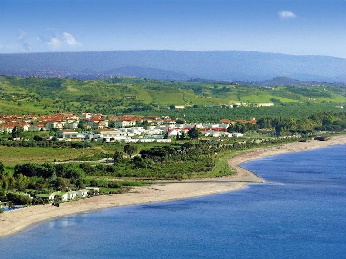 Luftaufnahme des Club Esse Sunbeach Village am Strand mit blauem Wasser und grünen Hügeln im Hintergrund.