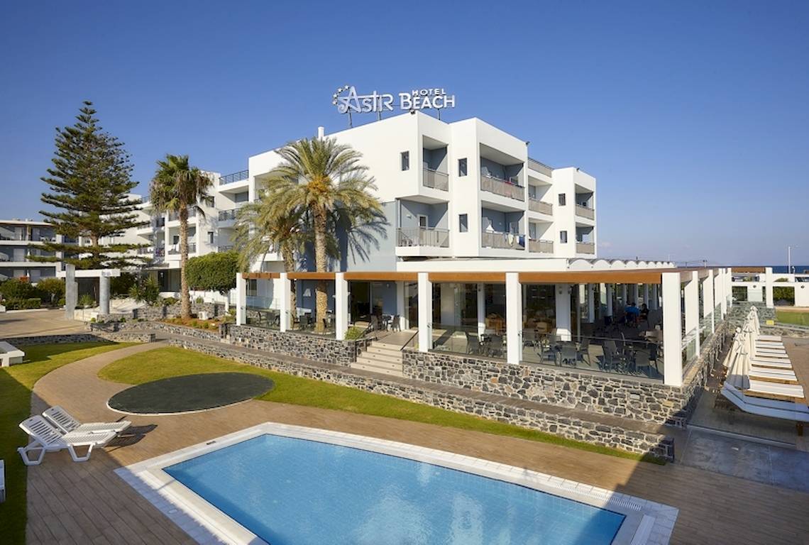 Außenansicht des Astir Beach Hotels auf Kreta mit Swimmingpool, Sonnenliegen und Palmen.