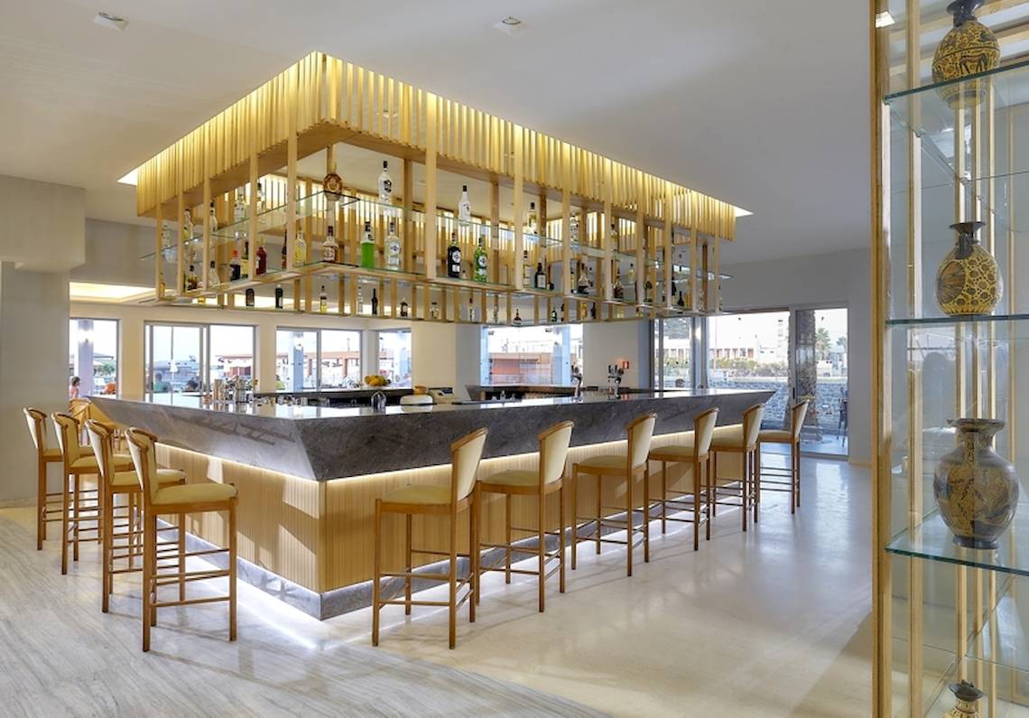 Bar im Astir Beach Hotel auf Kreta mit einer Reihe von Barhockern und einer Theke mit einer Glasvitrine oben.