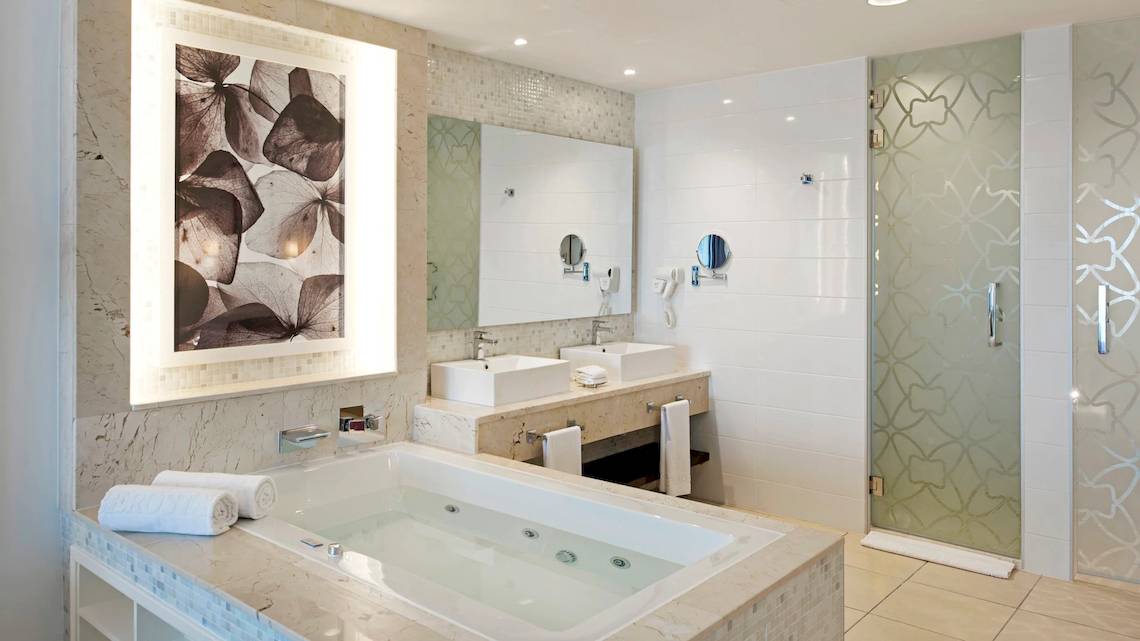 Ein Badezimmer im Iberostar Origin Bella Vista Varadero Hotel mit einer Badewanne, zwei Waschbecken, einer Glasdusche und einem Kunstwerk.