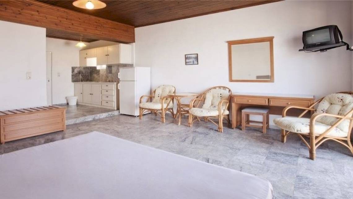 Ein modernes Schlafzimmer im Dedalos Beach Hotel mit weißem Bett, Holzmöbeln und einer Küche im Hintergrund.