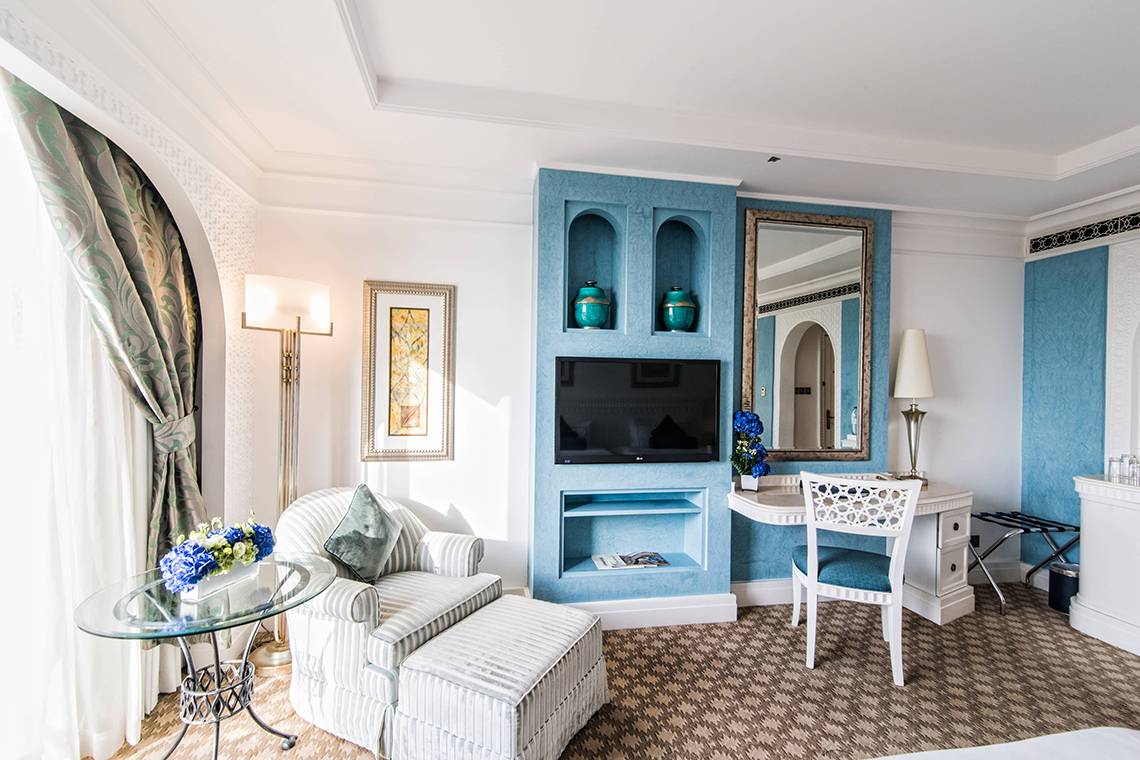 Innenansicht eines blau dekorierten Zimmers im Habtoor Grand Resort & Spa mit Fernseher, Schreibtisch und Stühlen.