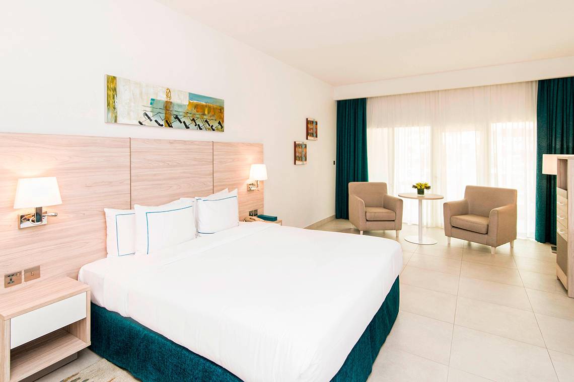 Ein elegantes Hotelzimmer im Habtoor Grand Resort & Spa mit einem Doppelbett, zwei Sesseln, einem Tisch und einem Kunstwerk an der Wand.