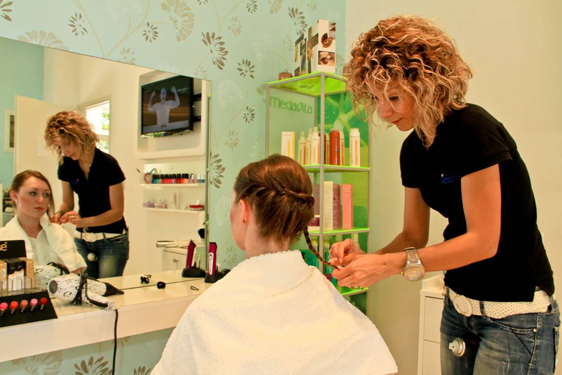 Friseur in einem Salon, der eine Kundin bedient, während eine weitere Kundin im Spiegel wartet.