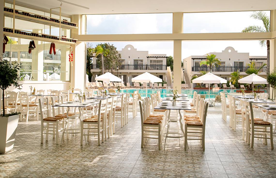 Poolseitiges Restaurant im Louis Paphos Breeze Hotel mit weißen Tischen und Stühlen unter Sonnenschirmen.