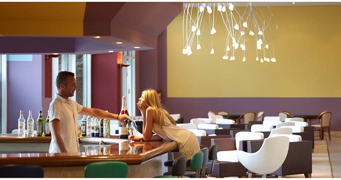 Ein Mann und eine Frau an der Bar im Esperides Beach Resort, beide mit Getränken, das Hotel im Hintergrund.