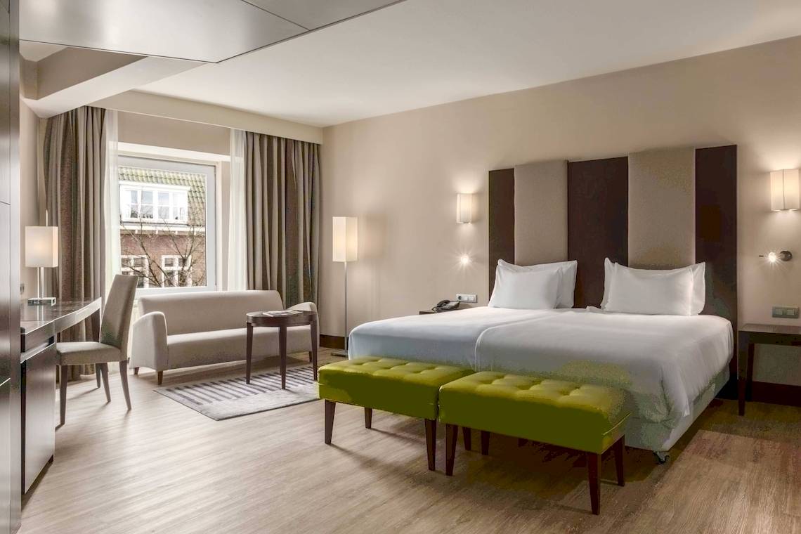 Modernes Zimmer im NH Amsterdam Centre Hotel mit zwei Betten, grünen Hockern und einem Fenster.