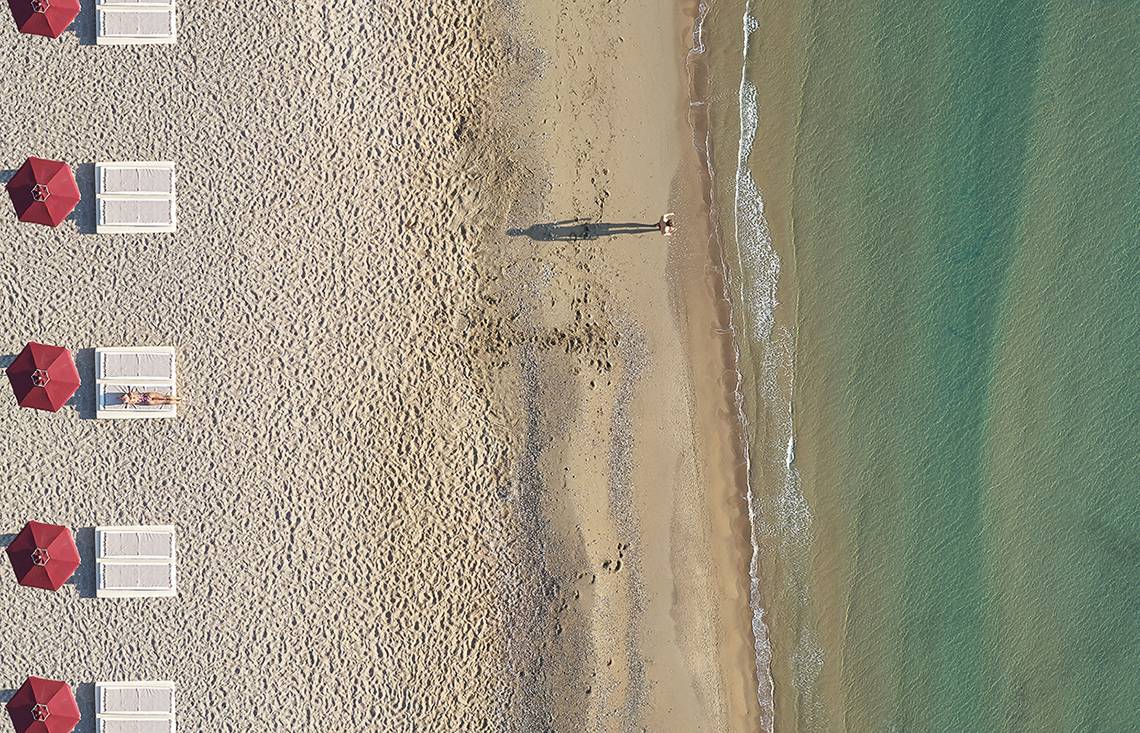 Luftaufnahme eines Luxushotels am Strand mit roten Sonnenschirmen und einer Person, die ins Wasser geht.