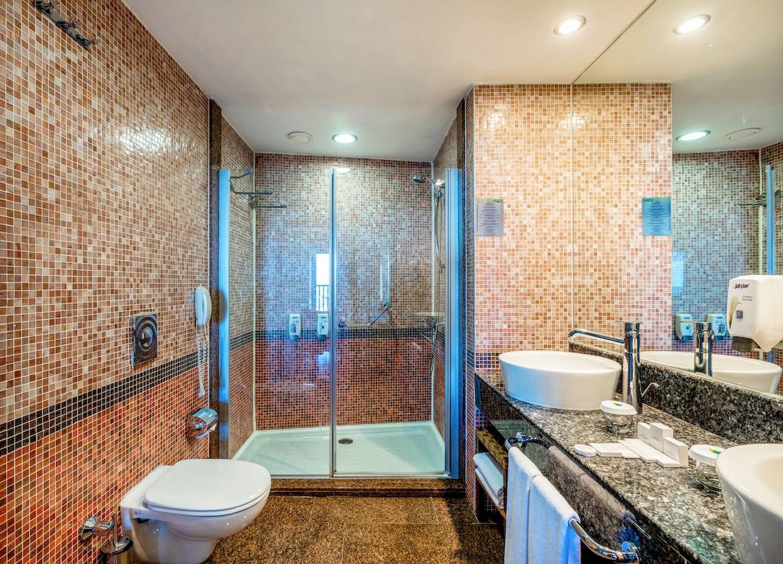 Ein Badezimmer mit einer Glasdusche, zwei Waschbecken, einem Spiegel und einem Toilettensitz im Asteria Family Resort Belek.