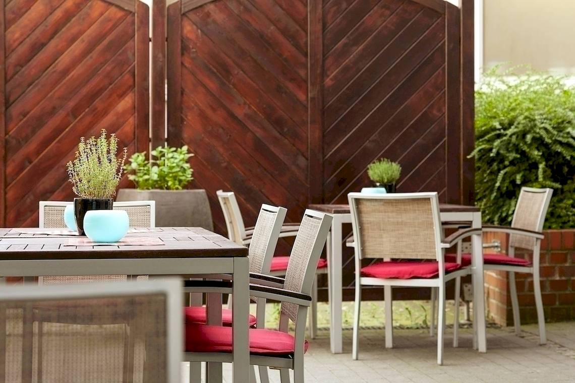 Terrasse des NH Hamburg Altona Hotels mit Tischen und Stühlen, Pflanzen und Holzwänden.