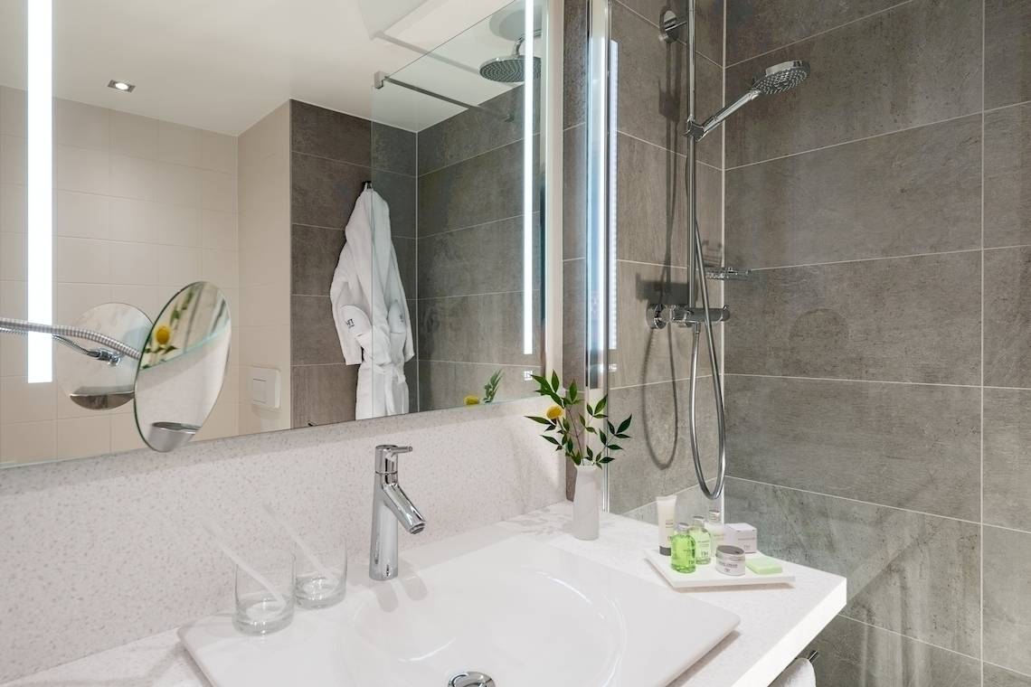 Badezimmer im NH Hamburg Altona Hotel mit grauen Fliesen, Spiegel und Handtuch.