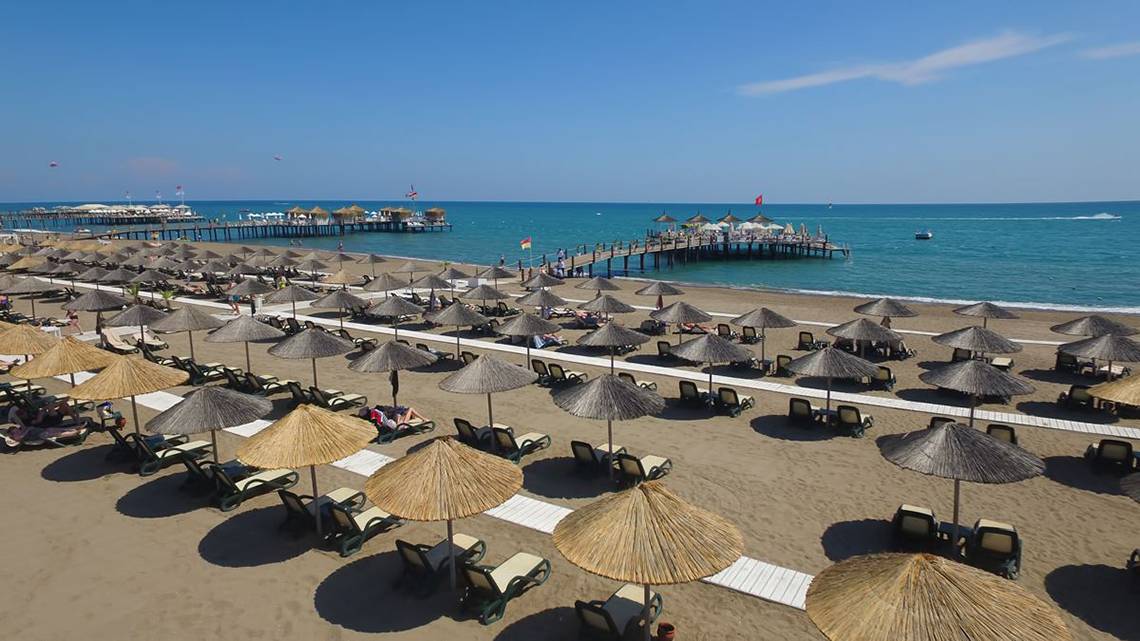 Strand des Baia Lara Hotels mit vielen Sonnenliegen und Sonnenschirmen, ein Pier im Hintergrund.