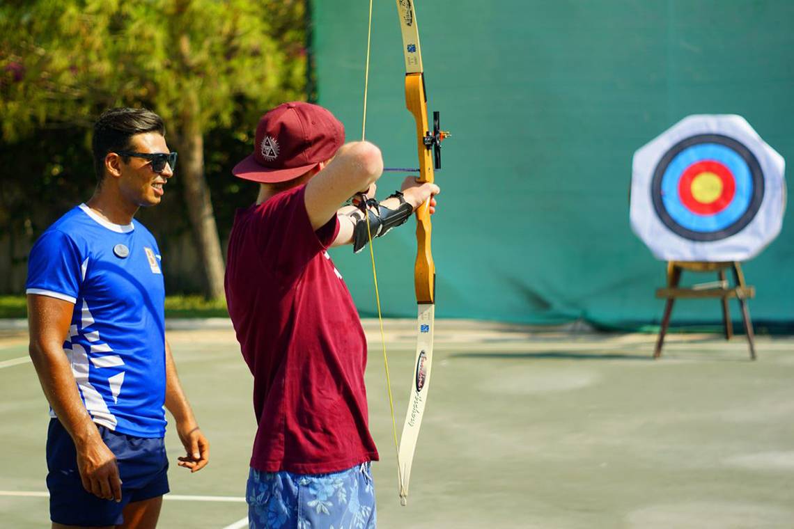 Ein Mann in einem roten T-Shirt und einer roten Kappe übt Bogenschießen auf einem Ziel auf einem Tennisplatz.
