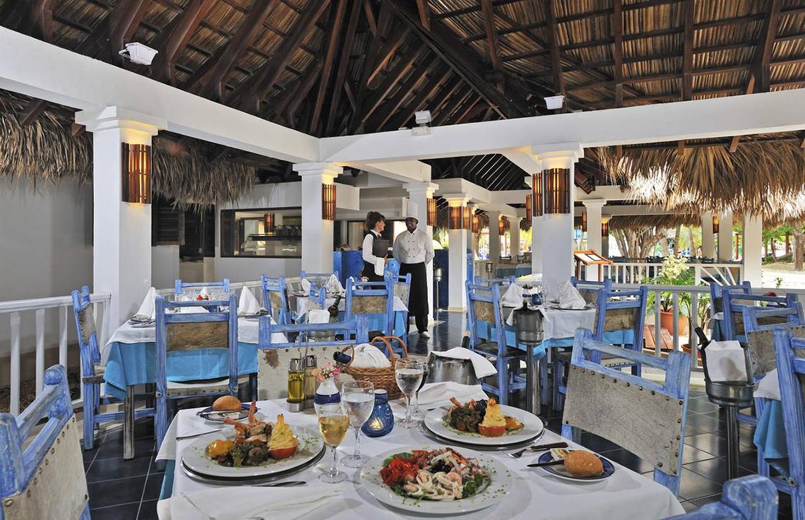 Restaurant im Sol Palmeras Hotel mit blauen Stühlen und Tischen, serviertem Essen und zwei Mitarbeitern im Hintergrund.