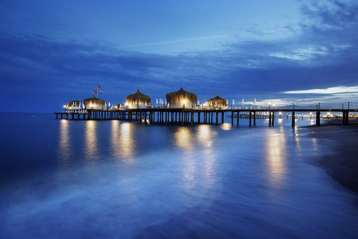 Abendlicher Blick auf die Pier des Liberty Hotels Lara mit leuchtenden Lampen und blauem Himmel.