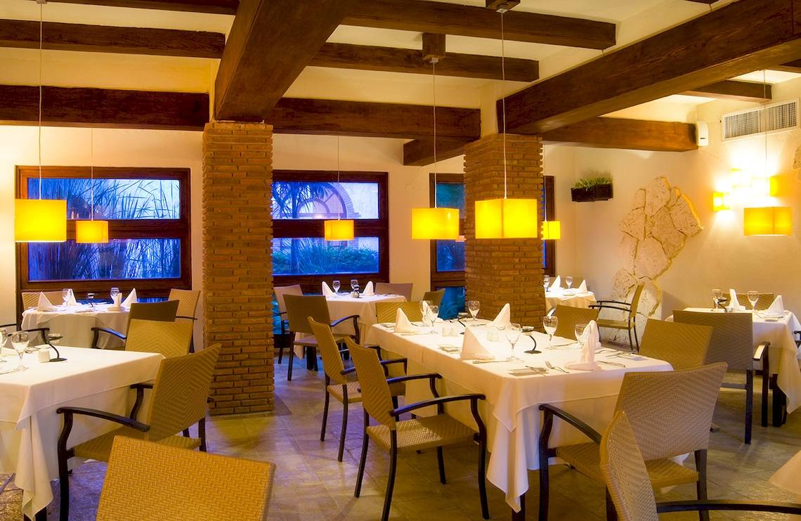 Innenansicht des Restaurants im IFA Villas Bavaro Hotel mit Tisch und Stühlen, hängenden Lampen und Fenstern.