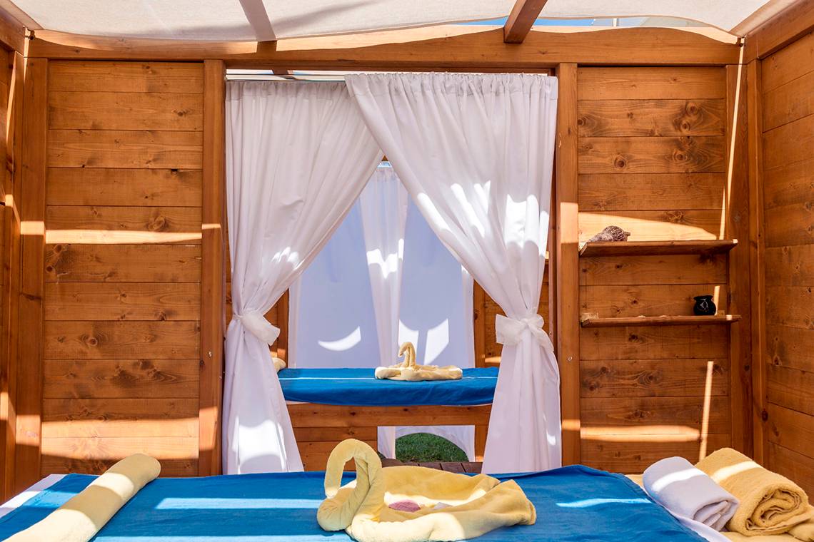 Massageraum im Civitel Creta Beach Hotel mit Holzwänden, weißen Vorhängen und blauen Liegen.