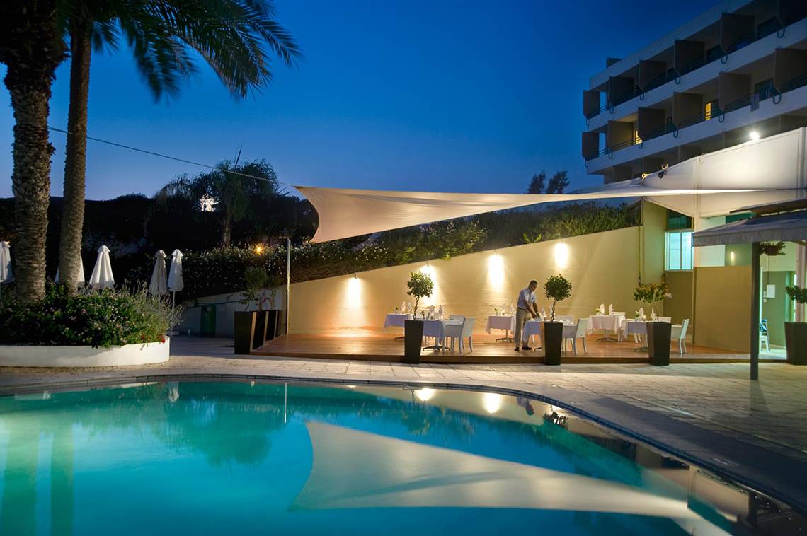 Abendlicher Poolbereich im Louis Imperial Beach Hotel mit Tisch, Stühlen und Palmen.