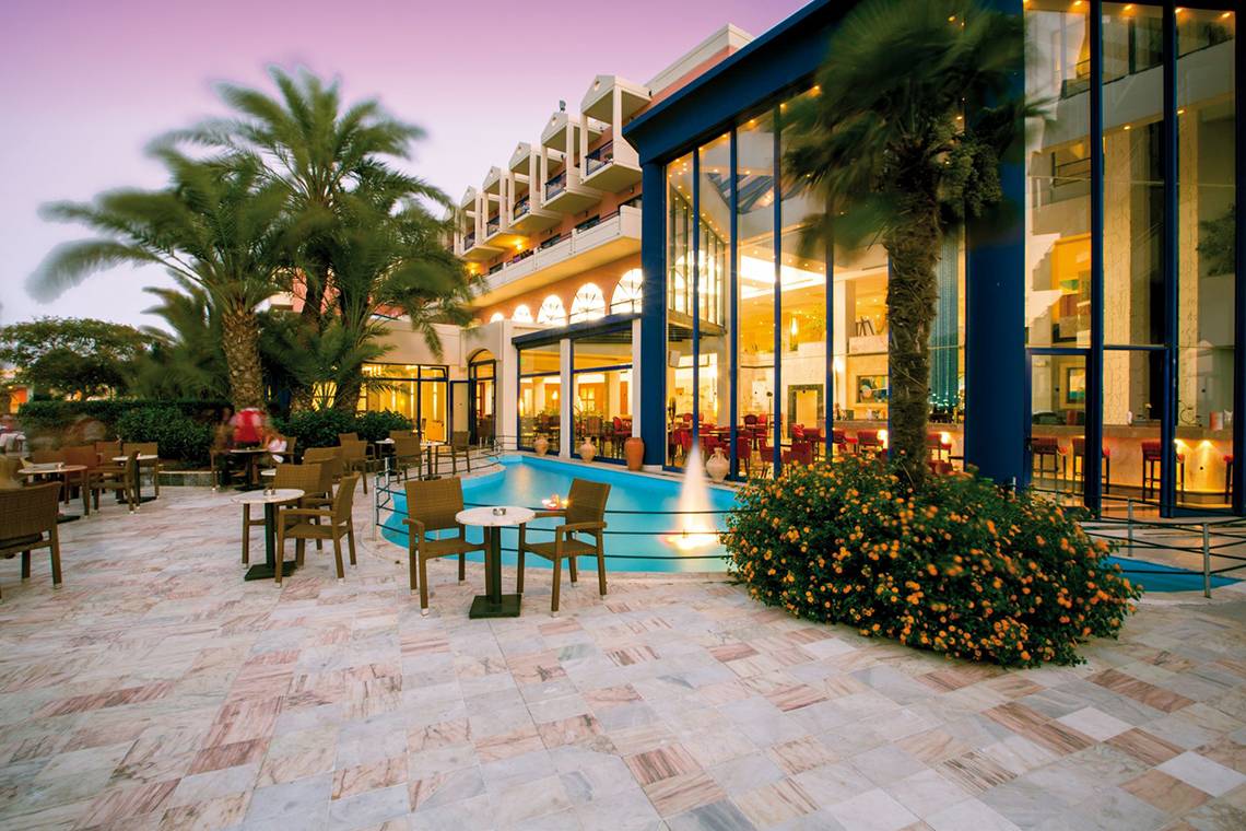 Außenbereich des Kresten Palace Hotels mit Swimmingpool, Tischen und Stühlen, umgeben von Pflanzen und Bäumen.